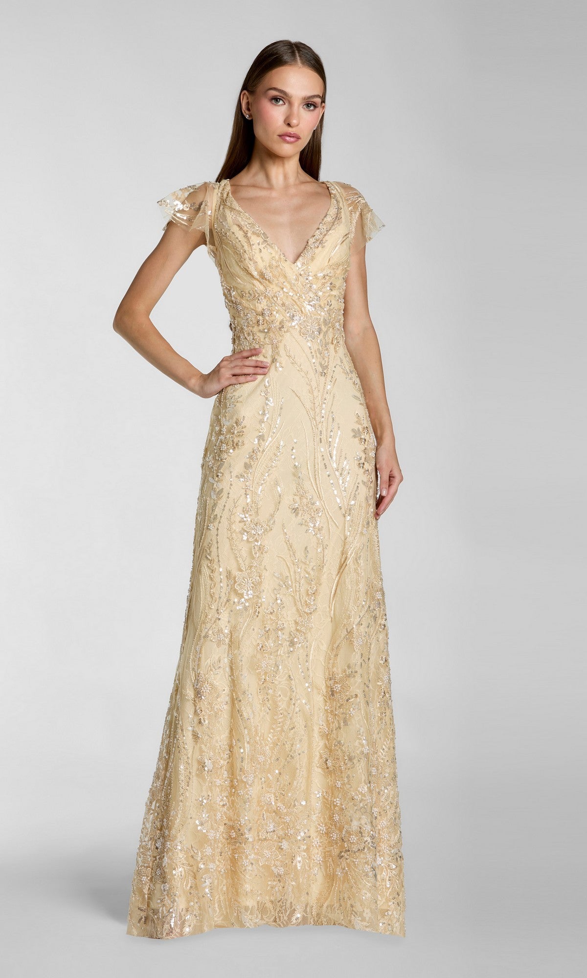 A woman stands against a plain light gray background, wearing the Long Formal Dress: Mac Duggal 68914 in gold—a floor-length, elegant gown adorned with sequins, featuring a V-neckline and short sheer sleeves.